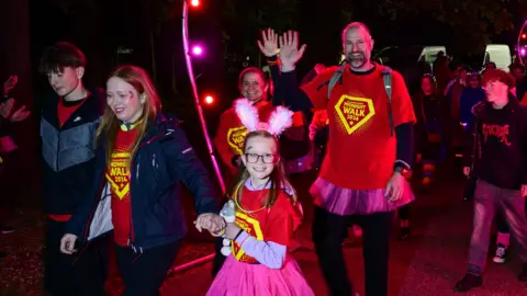 Nick Butcher Photography/St Elizabeth Hospice A family wearing matching red T-shirts walk in a large crowd of people during a charity event. They smile and some of the adults wave at the camera. 