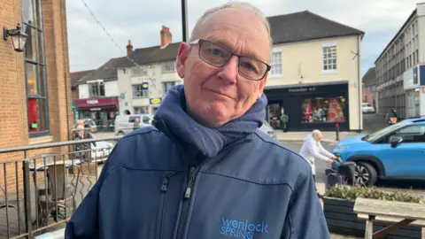 Stephen looking down the camera; he's wearing a navy coat with 'Wenlock Spring' written on it and a navy scarf. Behind him you can see cars, people, and shops decorated for Christmas on Newport's high street.