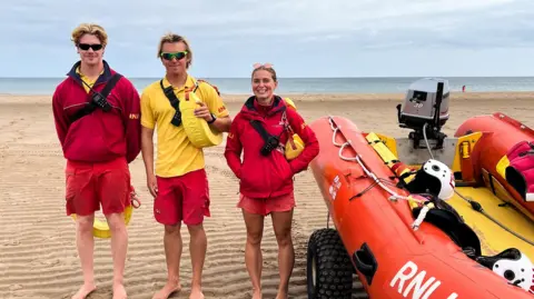 Three lifeguards standing in a line dressed in red and yellow next to a rescue boat. The one on the left has blonde floppy hair and is wearing black sunglasses. Matthew is standing in the middle with blonde hair and sports sunglasses, holding a floating device. A girl with her brown hair tied back is on the right.