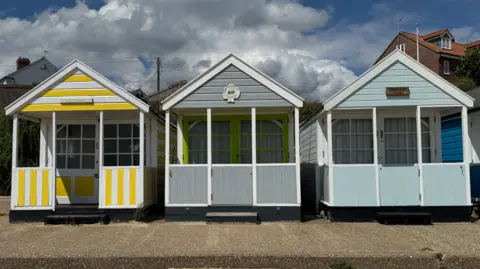 Three beach huts in a row including the middle one which is up for sale.  The lefthand side hut is yellow and white, the middle one grey and the righthand side one is pale mint blue.