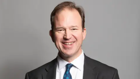 A portrait photograph of MP Jesse Norman. He is wearing a grey suit jacket and white shirt with a blue spotty tie. He is stood against a grey backdrop.