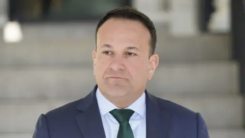Leo Varadkar has short dark hair. He is looking off to the side of the camera. He is wearing a blue suit jackers, a white shirt and a green tie.