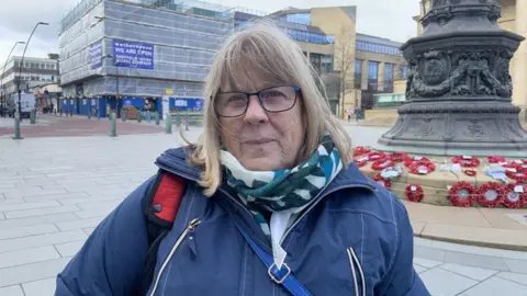 A woman with shoulder length blond hair is wearing glasses, a scarf and a blue coat. She is standing in front of a war memorial with poppy wreaths