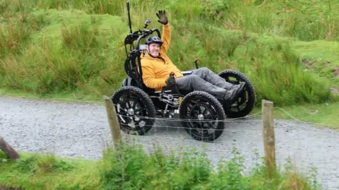 Nick Wilson Nick Wilson smiles and waves at the camera. He is wearing a yellow hoodie and grey trousers sitting in his specially designed chair. It has four big wheels and two steering handles. It has a black structure protecting Wilson's back and his head with a go pro positioned on the top. He is wearing a black helmet and gloves. He is outside with greenery surrounding him.