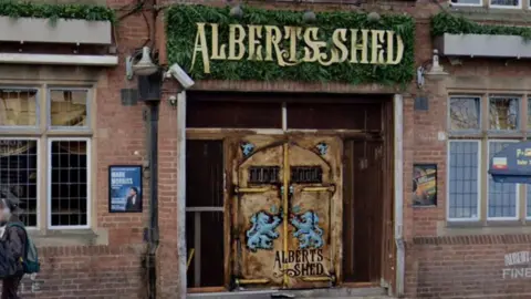 Google The entrance to a brick building, with the words Albert's Shed in gold lettering on a leafy green sign above a decorated wooden door that also reads Albert's Shed.