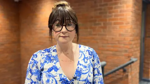 Harriet Crawford-Turner is wearing a v neck blue and white floral pattern dress though only her head and shoulders are visible in the shot. She is wearing dark-framed glasses and her dark hair is pulled back but her fringe and shorter lengths of hair are fraiming her face. She is stood at the top of a set of stairs on a landing. The grey railings of the stairs are partially visible in the background as are the red brick walls of the staircase.