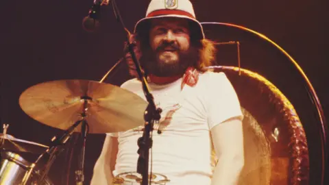John Bonham standing next to a set of drums. He is smiling at the camera and has dark brown hair and a beard. He is wearing a white t-shirt, red neck tie and hat which had a red band on it. 