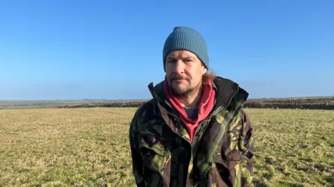 Dai Williams - operator of a pop-up campsite, stands in a camo jacket on a field wearing a teal woolly hat.