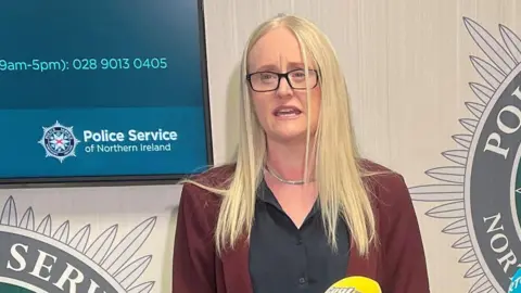 PA Media Geraldine Hanna speaking into microphones at a media conference. She is wearing a black blouse and burgundy jacket. She has long blonde hair and is wearing glasses.