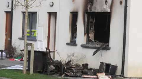 Two burnt out windows sit at the end of a row of three white terrace houses. There is black rubble and burnt household objects sitting outside on the green grass at the foot of the window. A single leafless tree sits in front of the objects on the grass. The other two houses are intact.