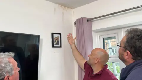 BBC Kevin stands in a room gesturing with his extended arm to the corner of a room where two walls meet the ceiling. There are brown marks on the white walls that appear to show the ingress of water. Two surveyors are standing either side of him, they are observing the marks on the walls and ceiling