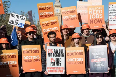 Getty Images A group of resident doctors stand with orange signs saying 'doctors need jobs' and 'fund the NHS' outside parliament in London on Tuesday