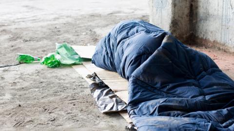 A sleeping bag with a person hidden from view inside on the street beside a building..