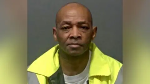Carson Grimes is clean shaven and wearing a high-vis yellow jacket as he stares at the camera in his police custody photograph