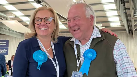 A woman with blonde hair and glasses in a blue top with a blue rosette has her arm around a man with grey receding hair in a green top and checked shirt also with a blue rosette.
