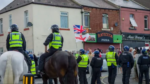 Ian Forsyth/Getty Images Mounted police and officers wearing riot gear line a street while protesters march through the high street waving Union Flags.