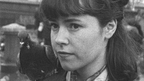 Black and white image of young female interviewee looking towards the camera with a pigeon behind her shoulder