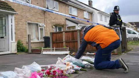 PA Media A man in an orange coat leaving a floral tribute outside the property on Bedale Drive, Leicester, where Chamiah Brindley died after a house fire.