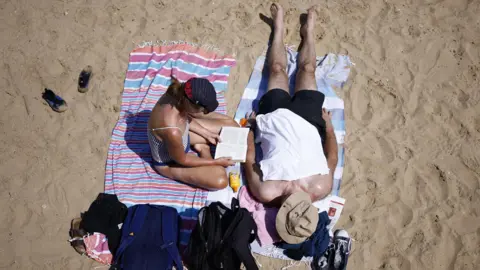 Two people sat on a sandy beach. A woman is sat up reading while a man is led out on a towel next to her. Both of them are wearing hats.