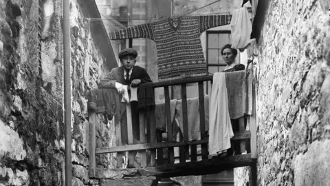 Shetland Museum & Archives A young boy and his mother standing in a closs, with their washing and a fair isle tunic jumper drying on a board.