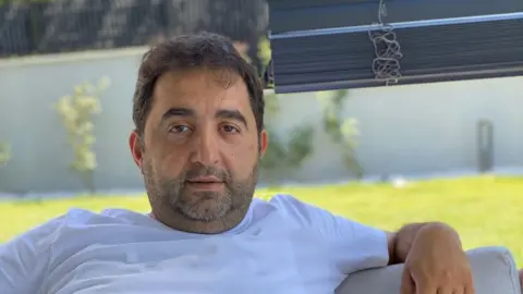Adem Savas, who is sat down looking at the camera while wearing a white T-shirt and with green grass illuminated by sunlight in the background.