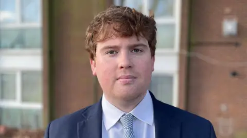 Dan Swords, who has medium length, light brown hair. He is standing in front of a block of flats with a serious expression on his face. He is wearing a navy suit jacket, white shirt and light blue tie.