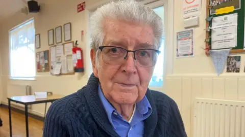 Tony Wadsworth. He has short white hair and glasses. He is wearing a blue shirt under a navy blue knitted cardigan. He is looking at the camera and smiling. 