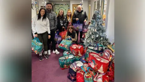 Samantha Taylor A group of four women and two men are holding large Christmas-themed bags and in front of them there is a large pile of bags. To the right of the photo, there is a decorated Christmas tree