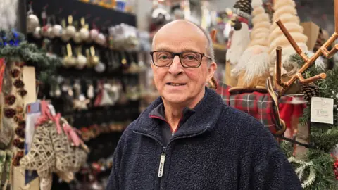 Paul Moseley/BBC Peter Hodges is wearing a dark navy fleece. He is standing in a shop in front of various Christmas-themed items such as Christmas tree decorations and toy santas.