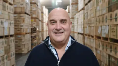 BBC Richard Clothier in his cheese storage facility, wearing a navy zip jumper and a light coloured top beneath and smiling at the camera. There are boxes of cheese behind him. 