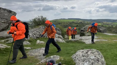Young people wearing orange waterproofs and orange woolly hats are carrying rucksacks and walking across granite outcrops and grass.