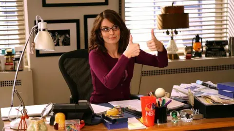 'AI can't write comedy', says Saturday Night Live star 7 NBC Photo Tina Fey as Liz Lemon in 30 Rock - wearing a stripy sweater ad glasses she sits at her desk on the show, giving a two-thumbs-up sign to someone off to the left.