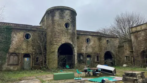 Photograph of the derelict stable block at Winstanley Hall in Wigan. The image shows exposed timbers and trees growing up the side of the building.
