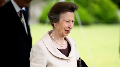 Princess Anne. She has dark grey hair and is wearing a cream jacket, a pearl necklace and pearl earrings. She is pictured outside, squinting in the sun and looking to the side. Two Korean men are stood behind her. 