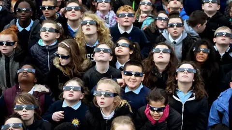 Getty Images School pupils all wearing eclipse glasses and looking up towards the sky. The group includes a mix of boys and girls and they are wearing a navy blue school uniform.