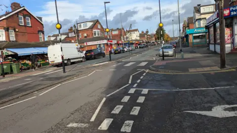 A main road in a city suburb. A zebra crossing, parked vehicles and shop fronts are all visible on both sides of the road. 