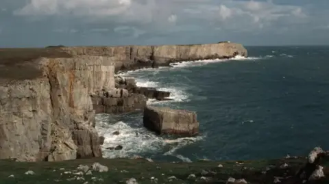 Getty Images Cliffs at St Govan's Head. The sky is blue and cloudy. The waves crash into the cliff face.