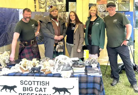 Scottish Big Cat Research Team A group of five people in a row. They are dressed in various types of Scottish country dress. Before them is a tartan-covered table with a variety of books and objects. There is a banner with two black big cats and the name Scottish Big Cat Research on it. Behind there are various displays. 