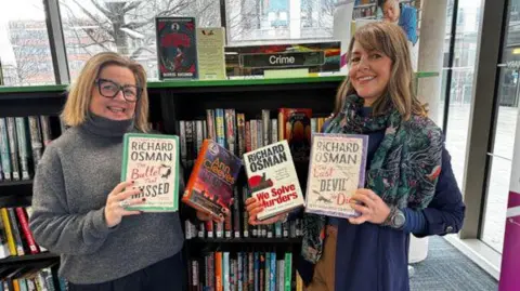 Two women stand next to each other and smile at the camera while holding a book in each hand. Three of the four books are written by Richard Osman and the fourth is by Ann Cleeves.