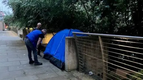 A stretch of pavement with a fence to one side, overhung with trees, and a blue tent belonging to a homeless person pitched in the middle. Two men are leaning over in a manner suggesting they are communicating with someone inside.