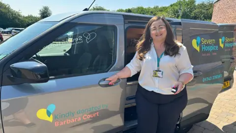 A woman with long brown hair is wearing a white shirt, black trousers and a blue lanyard. She is standing in front of a small grey van with yellow and blue lettering and logos on it. Writing says "Severn Hospice". On the passenger door it reads "Kindly donated by Barbara Court".