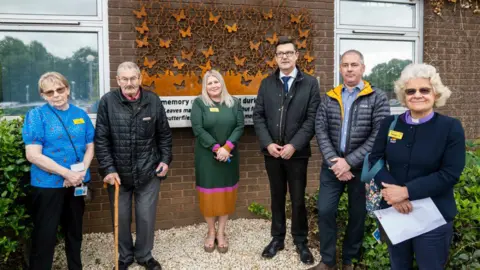 Worcestershire Acute Hospitals NHS Trust Staff standing in front of the steel memorial mounted on a wall, which has butterflies and a quote