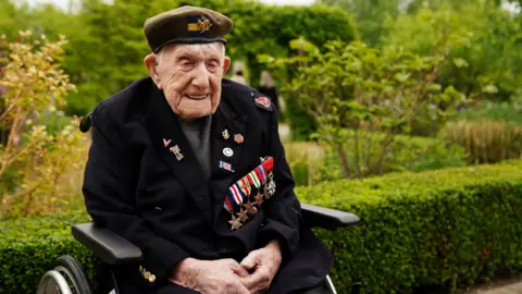 PA Media A veteran wearing a beret and a number of medals on a blue jacket, sits smiling in a wheelchair.