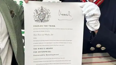 Green Doors A close up of the certificate from King Charles III, complete with signature and his coat of arms. It is being partly held up by Susan Lousada, the Lord-Lieutenant of Bedfordshire, who is wearing white gloves.