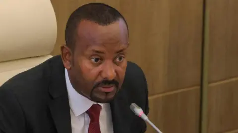 Head and shoulders shot of Abiy Ahmed in a red tie and dark jacket speaking into a microphone. 