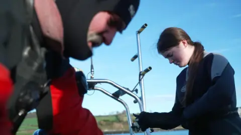 A man wearing a red and black diving suit, he is looking down he has a grey moustache. Beside him is a girl wearing a black and grey diving suit, she has dark hair, she is looking down.