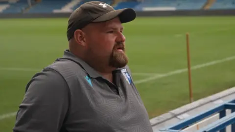 A man with a baseball cap and goatee beard standing next to a football pitch. 