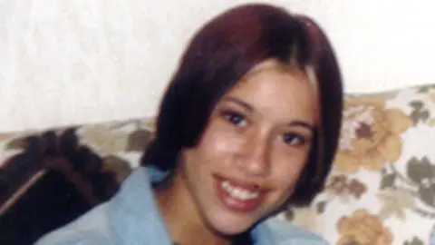 Suffolk Police A slightly blurred image of Tania Nicol taken before her murder in 2006. She has short dark red hair and is slightly tilting her head down to the right as she smiles at the camera. She is wearing a blue top with a collar on it. She is sitting on a floral sofa.