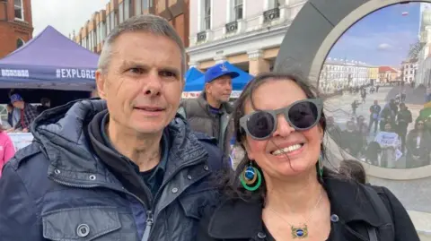 Neil and Ana Ball are looking at the camera from Ipswich town centre, in front of the "portal" to Brazil. She has dark hair which is tied back and sunglasses on. He has short, grey hair and is wearing a blue top and jacket.