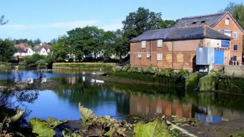 Historical brick mill buildings are reflected in the calm water of the River Itchen on a sunny day. Trees and houses are the background.